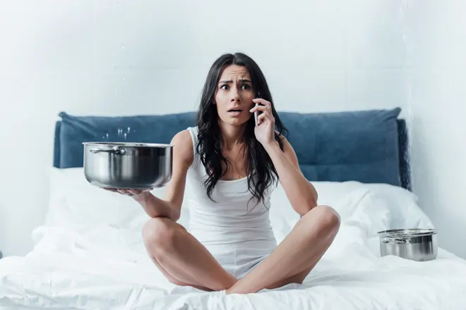 Shocked woman talking on smartphone and holding pot under leaking ceiling in bedroom Pinnacle Carpet Cleaning & Restorations florida